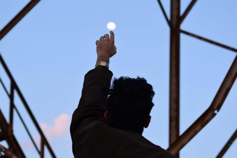 A man silhouetted against the morning sky reaches towards the moon with an outstretched hand.